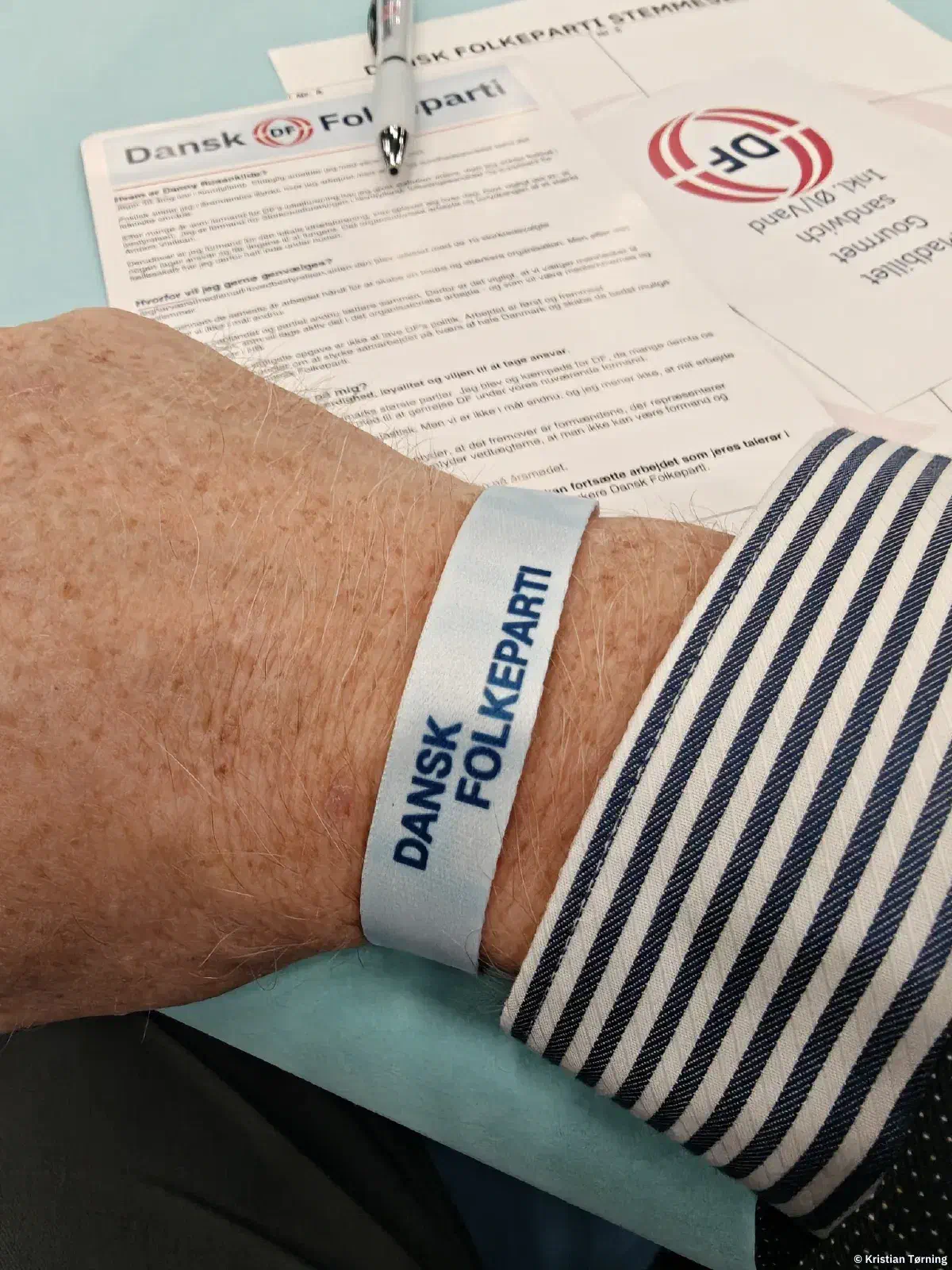 Håndled med et hvidt armbånd med teksten Dansk Folkeparti over mødepapirer