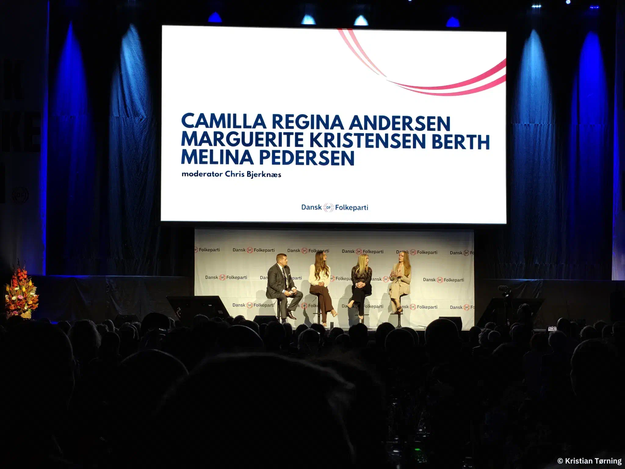 Fire personer i panelsamtale på scenen under en skærm med deltagernes navne