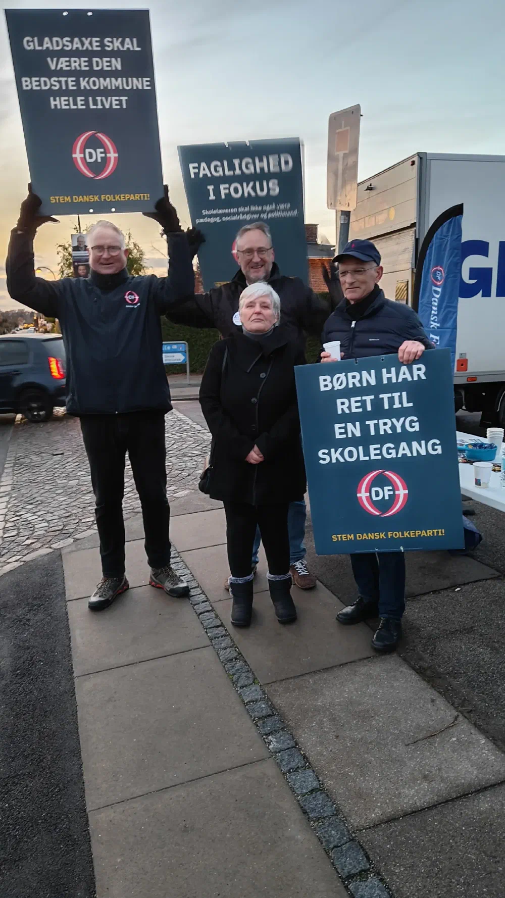 Fire personer holder DF-plakater om skole og tryghed i vejret på et gadehjørne